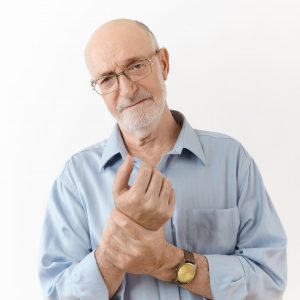 People, age, well being, illness and health problems concept. Studio shot of frustrated upset sixty year old man in spectacles having painful look, rubbing wrist, suffering from pain in joints